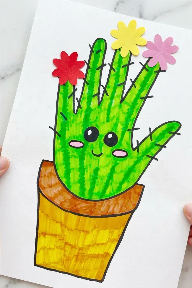 A hand-drawn craft showing a smiling green handprint transformed into a cactus in a yellow-brown pot. It's decorated with tiny black spines and three colorful paper flowers (red, yellow, and pink) on top, all on a white background.