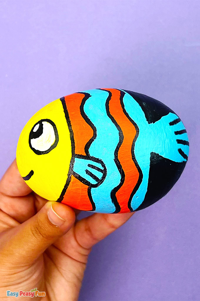 A colorful fish painted rock with orange and blue stripes, a yellow head, and a smiling face held in a hand.