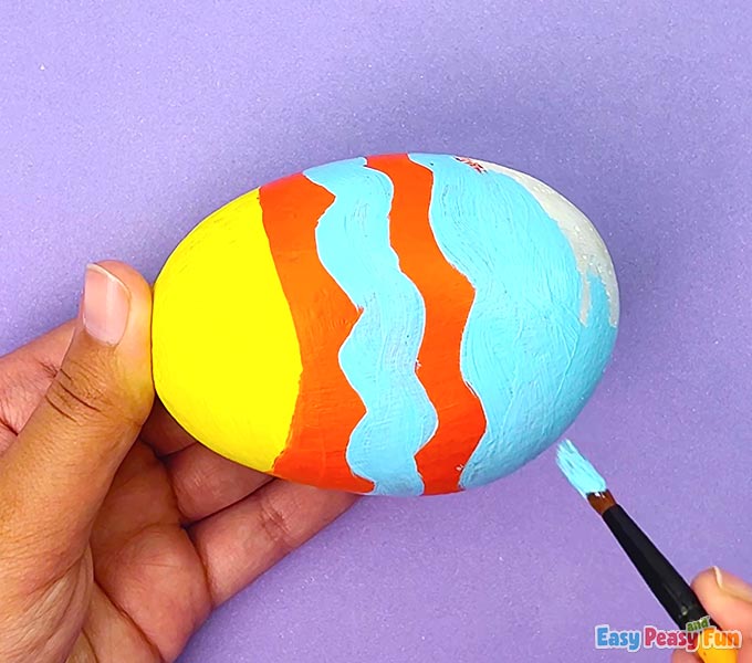 Adding light blue wavy stripes and a small fin to an orange and yellow painted fish rock.