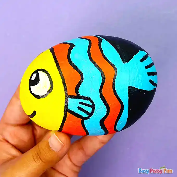 A colorful fish painted rock with orange and blue stripes, a yellow head, and a smiling face held in a hand.