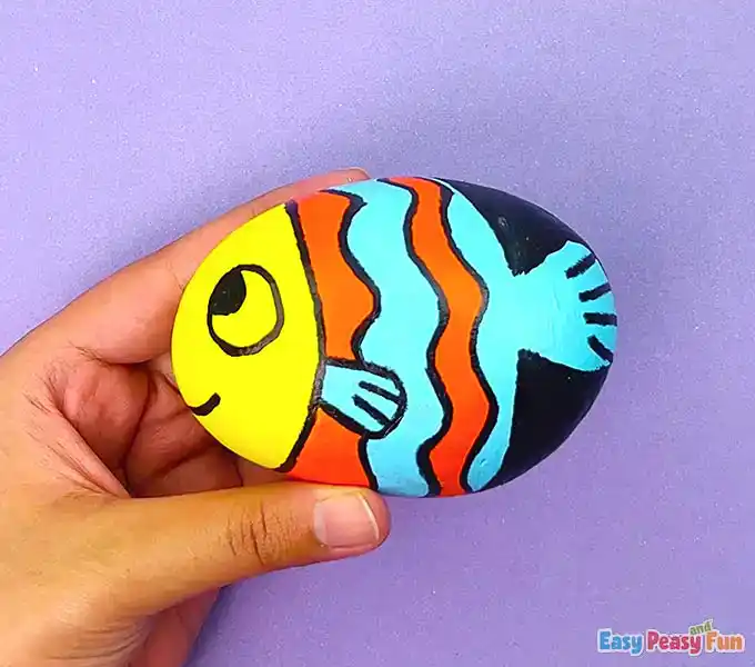 Using a black marker to outline blue patterns and draw a smiling mouth on a painted fish rock.