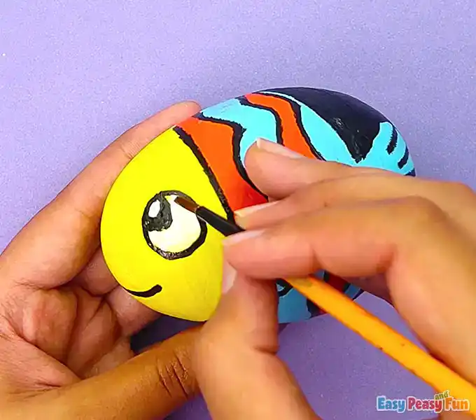 A close-up of a hand adding a tiny white highlight to the eye of a painted fish rock for a realistic finish.