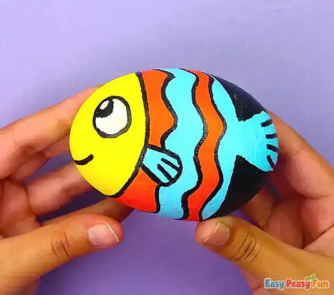 A colorful fish painted rock with orange and blue stripes, a yellow head, and a smiling face held in a hand.