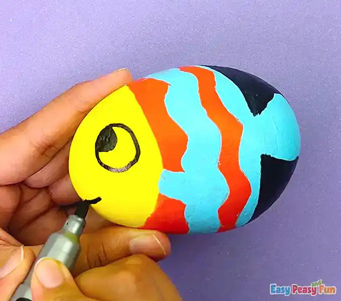 Using a black marker to outline blue patterns and draw a smiling mouth on a painted fish rock.