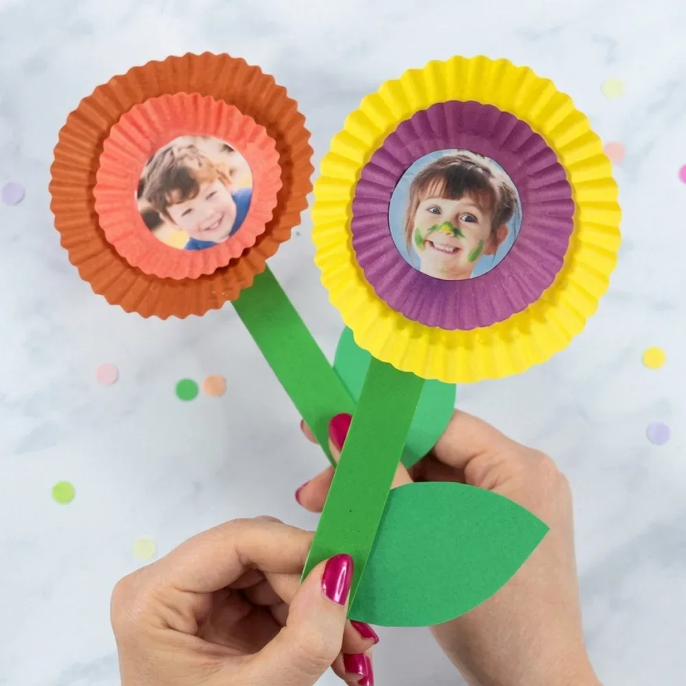 Two handmade paper flowers made from layered orange, yellow, and purple cupcake liners with circular children’s photos in the center, attached to green paper stems.