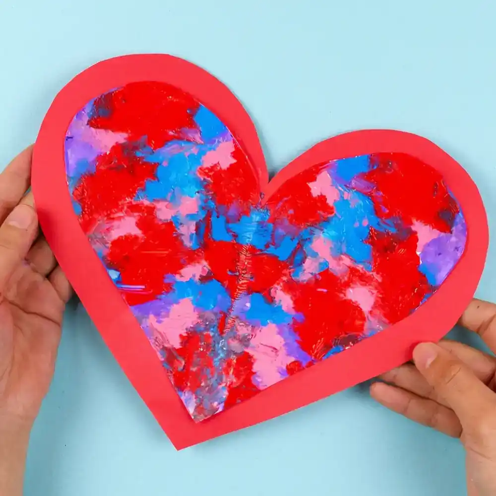 A person holding a large red paper heart featuring a colorful, symmetrical center created by a blue and red paint-smearing technique.