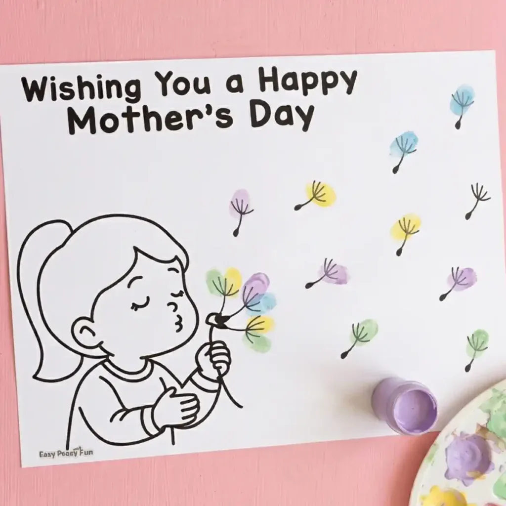 Mother's Day fingerprint craft featuring a girl blowing dandelion seeds, with colorful pastel fingerprints on the seeds, laying flat on a pink wooden table next to a paint palette.