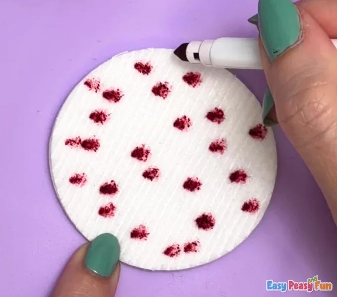 A hand holds a marker, carefully pressing the tip onto a white, textured cotton pad to create a scattered red dot pattern.