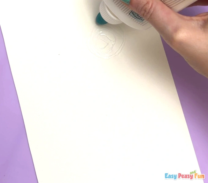 A hand is gently pressing the dried, colorful cotton pad onto the glue circle on the white paper to ensure it sticks well.