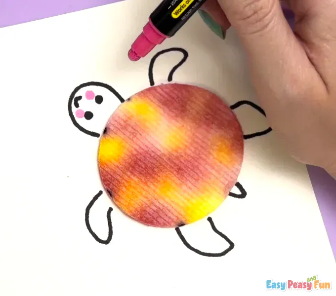 A pink marker is being used to color in two small, circular blushing cheeks on the turtle's face next to the eyes.