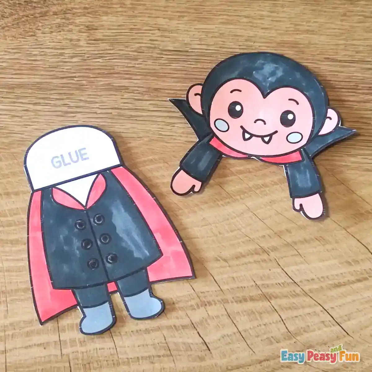 Two cut-out paper vampire pieces consisting of a body with a glue tab and a separate head with outstretched arms on a wooden surface.