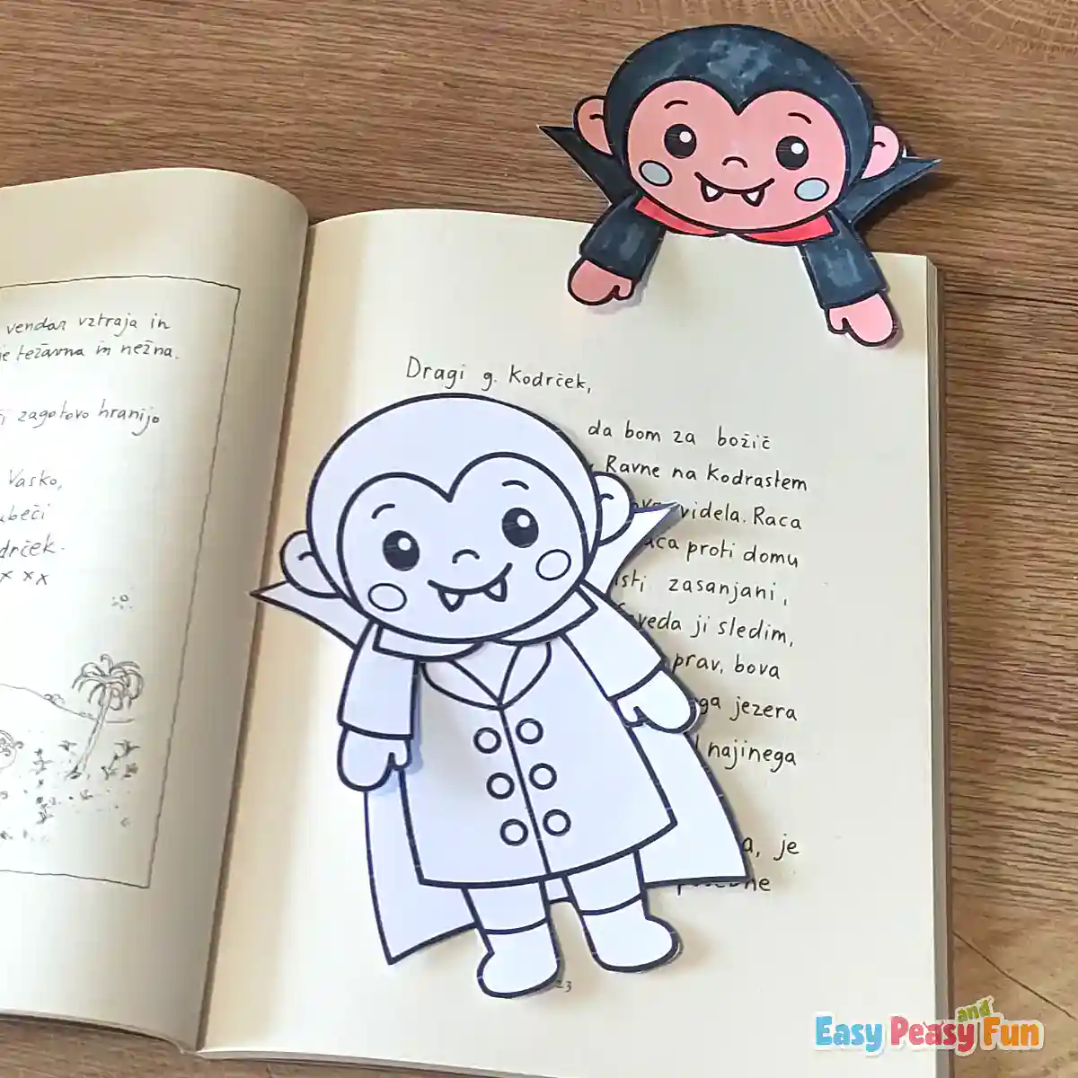 A completed vampire bookmark gripping the top corner of an open book page, with an uncolored version resting below.