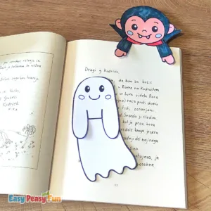 A colored vampire bookmark at the top of an open book, with an uncolored ghost template resting on the page below.
