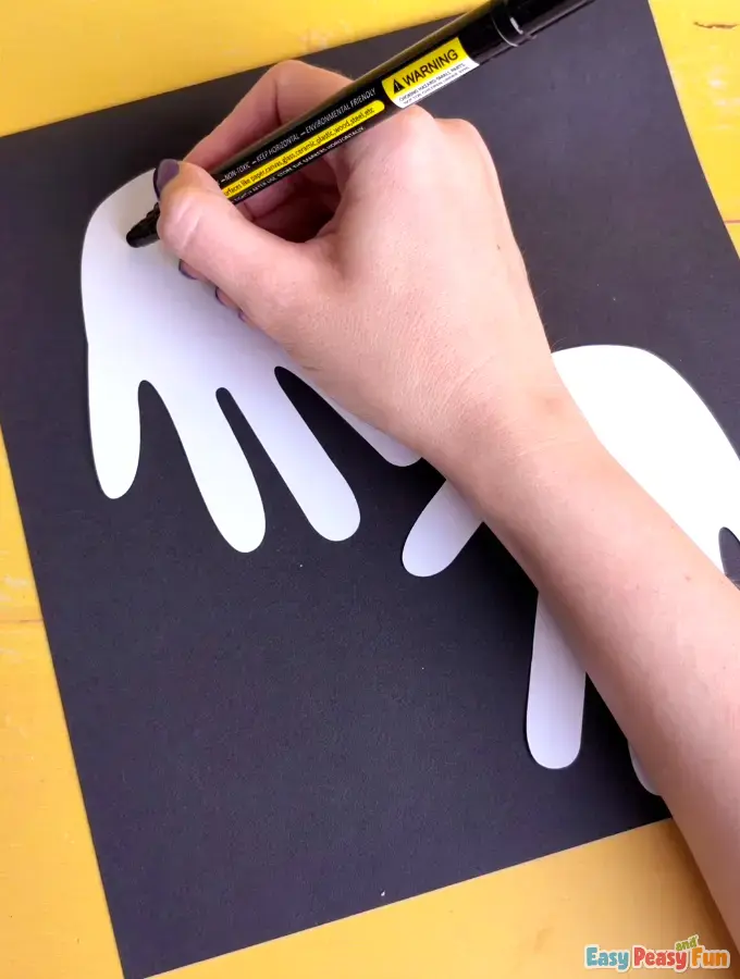A hand using a black marker to draw eyes and a mouth on a white handprint shape glued to black paper.