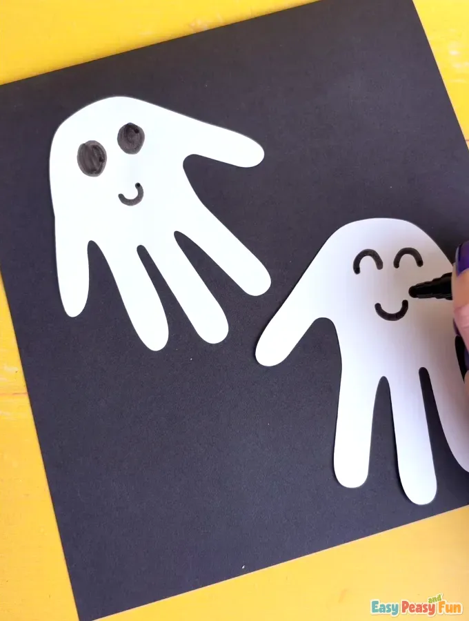 A hand drawing a smiling mouth on the second white handprint ghost shape with a black marker.