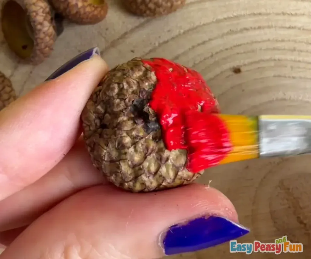 A paintbrush applies vibrant red acrylic paint across the top of the acorn cap.