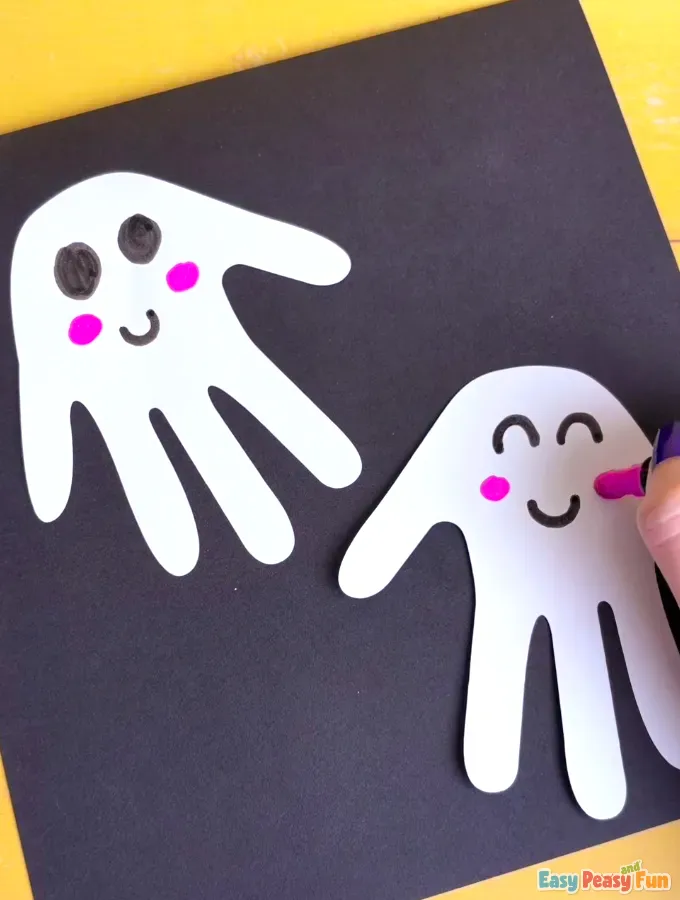 A hand using a pink marker to draw rosy pink cheeks on the white ghost handprint cutouts.