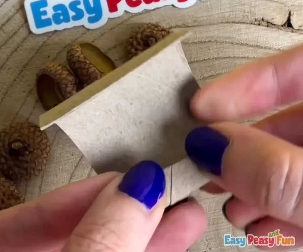 Fingers begin rolling and folding the cardboard strip to form a cylindrical shape.