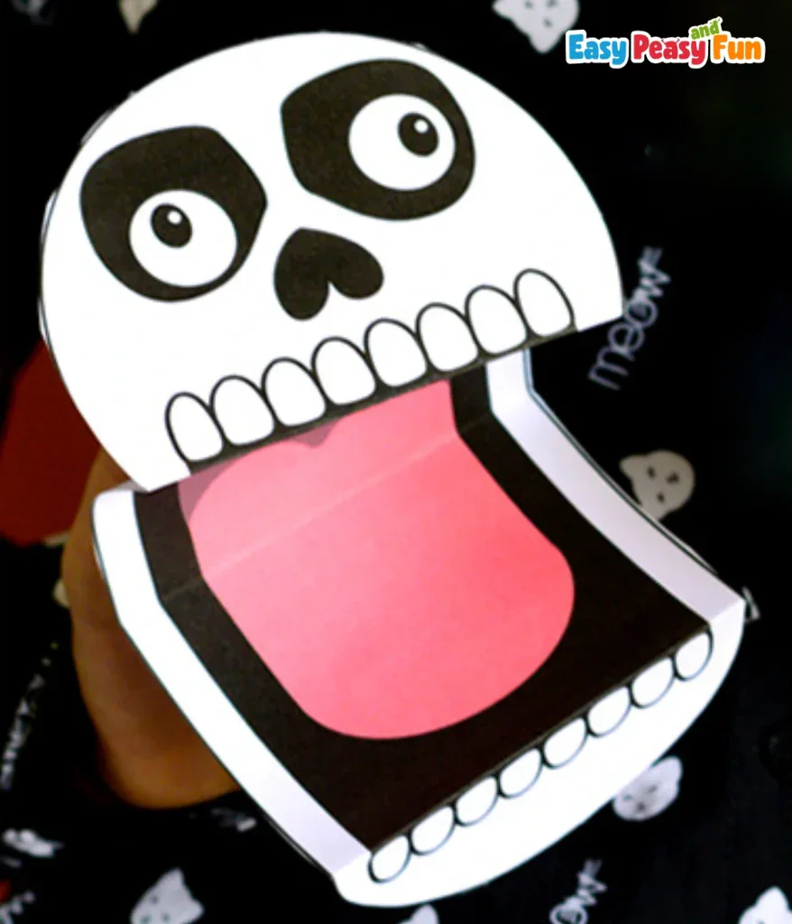 A hand holding a paper skeleton puppet with a wide-open mouth showing a pink tongue.